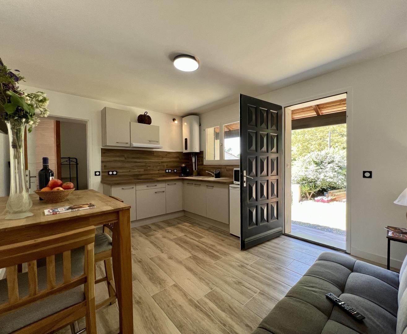 A modern kitchen with wooden cabinets, a dining table with a vase and fruit bowl, and a grey sofa with a remote control.