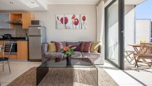 Un espacio de estar moderno con una mesa de centro de cristal, un sofá con cojines, una cocina equipada y un ventanal corredero que da a un área exterior.
