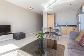 Een moderne woonkamer met een flatscreen televisie, een bijzettafeltje met een plant, een bank en een eethoek met een tafel en stoelen.