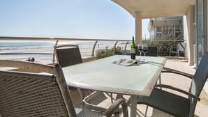 A balcony with a glass table, chairs, and a bottle of wine. The balcony overlooks a beach and the ocean.