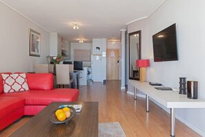 Una sala de estar moderna con un sofá rojo, una mesa de centro con naranjas, un televisor de pantalla plana y un área de comedor al fondo.
