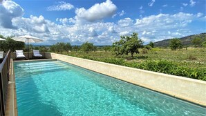 Una piscina rettangolare con acqua cristallina azzurra, circondata da un basso muro color crema. Sul lato sinistro, ci sono lettini bianchi e un ombrellone.