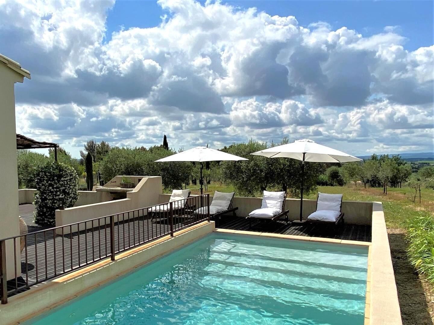 A pool area with lounge chairs and umbrellas, surrounded by greenery and a clear sky.