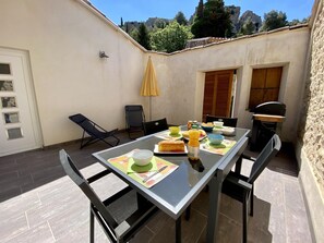 Een eettafel met stoelen, een gele parasol en een barbecue in een binnenplaats met uitzicht op rotsachtig terrein.