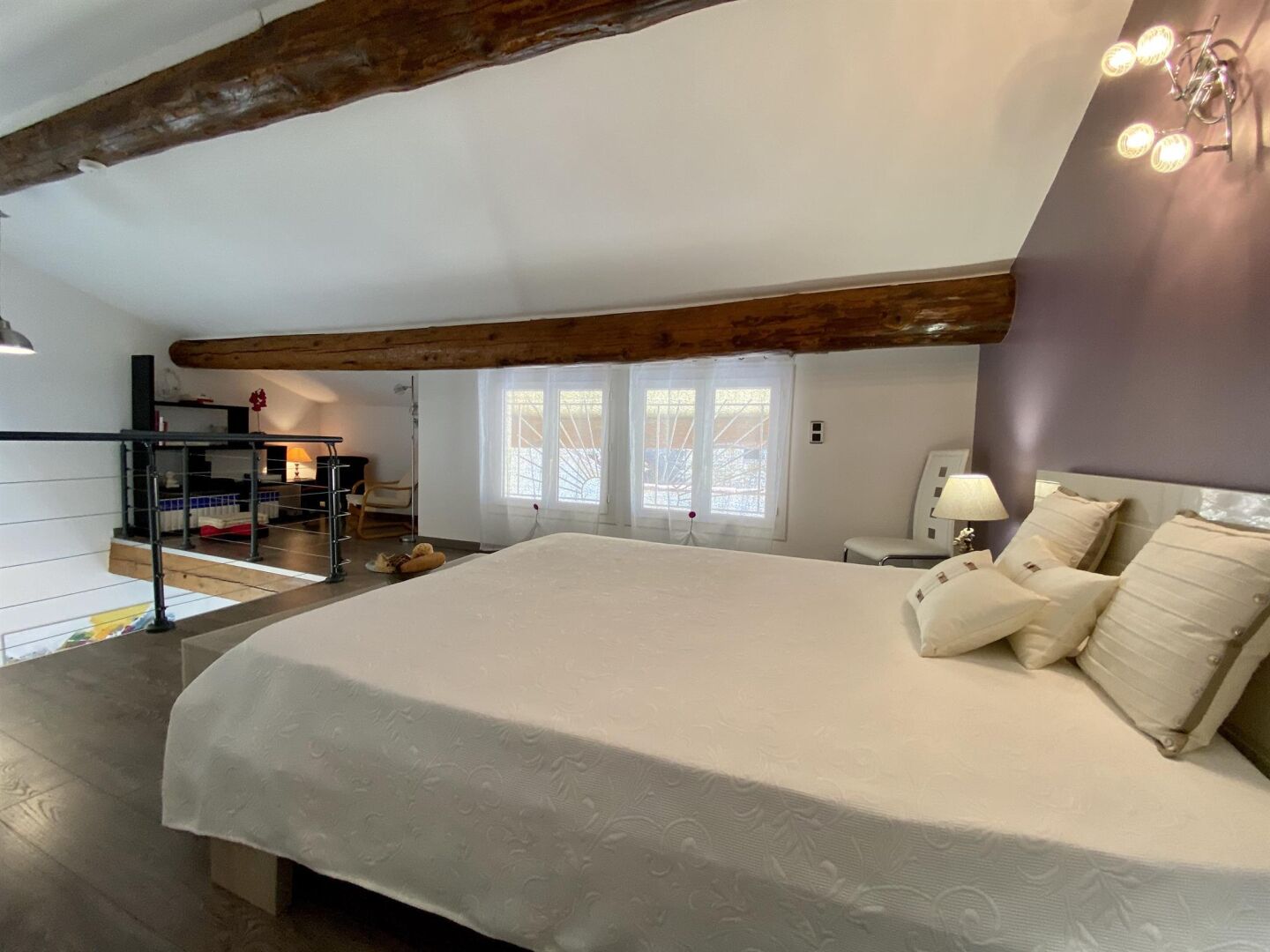 A bedroom with a large bed, a wooden ceiling beam, a window with a view, and a bedside table with a lamp.