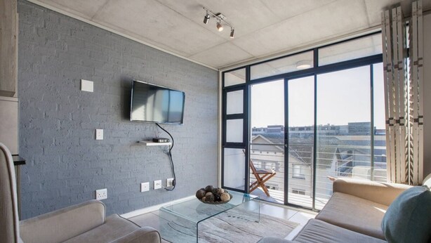 A modern living room with a large window, a flat-screen TV mounted on the wall, and a glass coffee table. - Key West 417 by Ctha (Milnerton)