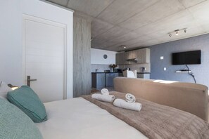A modern hotel room with a large bed, a flat-screen TV mounted on the wall, and a kitchenette in the background.