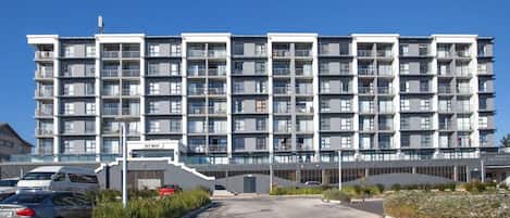 A multi-story residential building with balconies, a parking area, and a clear sky.