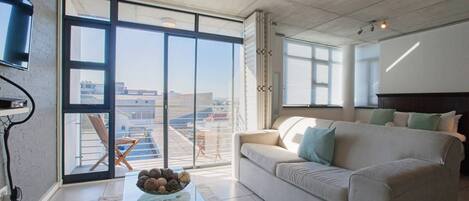 Amplia sala de estar con un gran ventanal corredero que da a una terraza exterior, un sofá con cojines, una mesa de centro con adornos y un televisor de pantalla plana montado en la pared.