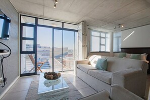 A spacious living room with a large sliding glass door leading to an outdoor terrace, a sofa with cushions, a coffee table with decorative items, and a flat-screen TV mounted on the wall.