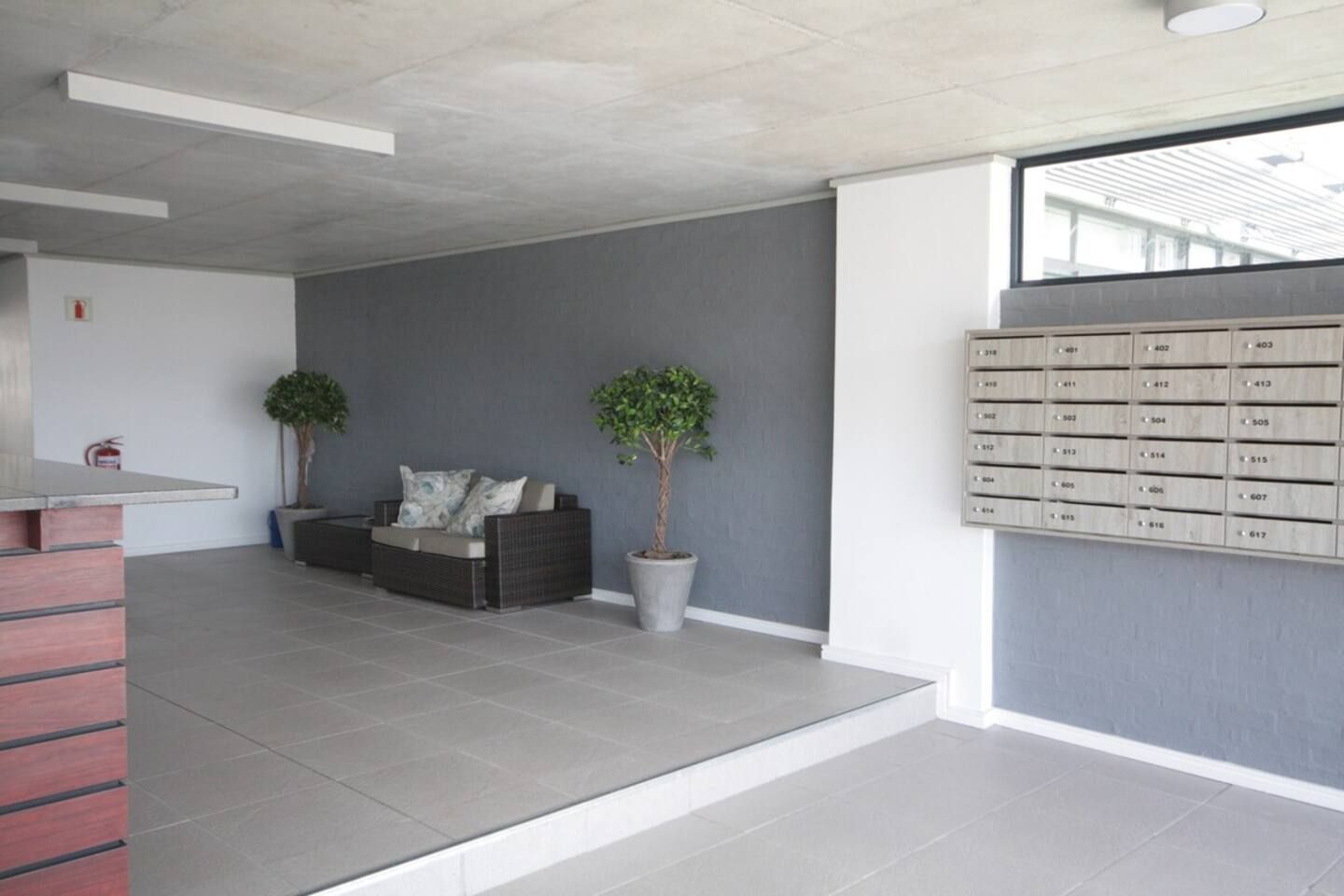Un interno moderno con un divano grigio, due piante in vaso, una reception e una cassetta postale a parete.