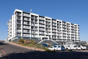 A multi-story building with a modern architectural design, featuring balconies and a parking area with several vehicles.