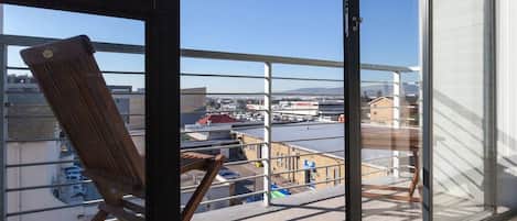 A balcony with a wooden chair, a glass door, and a view of a parking lot and buildings.