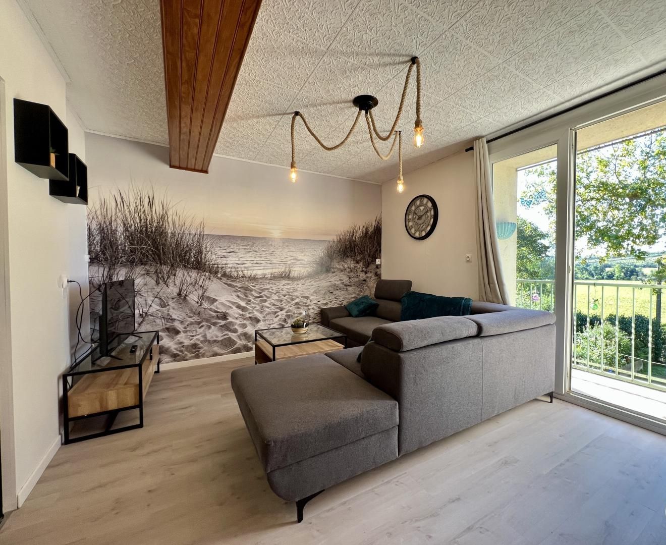 Una sala de estar moderna con un amplio sofá seccional gris, una mesa de centro y un televisor sobre un mueble bajo. Hay un ventanal con vistas a una playa y un balcón con barandilla.