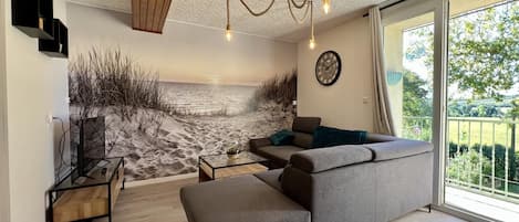 A modern living room with a large gray sectional sofa, a coffee table, and a TV on a stand. There is a large window with a view of a beach and a balcony with a railing.