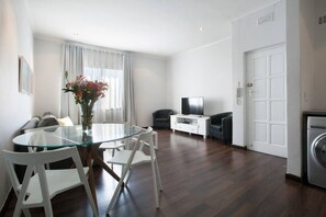 A modern living room with a glass dining table, white chairs, a sofa, a TV stand, and a washing machine.