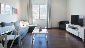 A modern living room with a sofa, a glass coffee table, a TV on a white cabinet, and a view through a large window. - Polana Court 12 by CTHA (Cape Town)