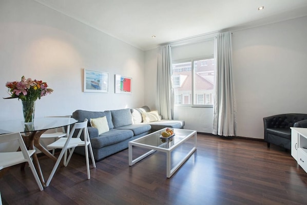 A modern living room with a glass coffee table, a grey sofa, and a dining table with chairs.