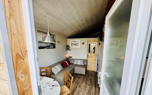 Een kleine leefruimte met een houten vloer, een witte tafel met bord en bestek, een bank met kussens en een houten deur.
