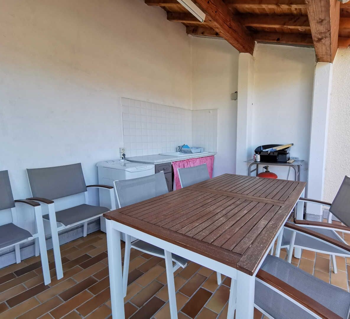 Een buitenpatio met een houten tafel en stoelen, een keukentje met een spoelbak en gasfornuis, en een betegelde vloer.