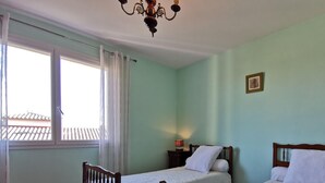A room with two single beds, a window with curtains, a chandelier, and a small table with a lamp.