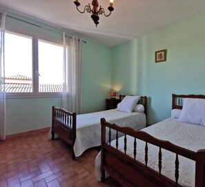 Una habitación con dos camas individuales, una ventana con cortinas, una araña y una mesita con lámpara.