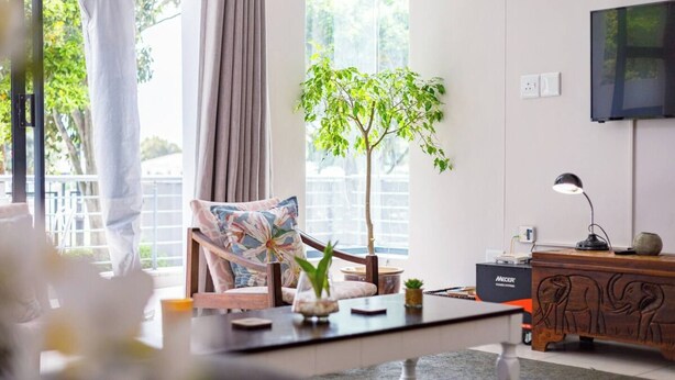 Una sala de estar moderna con un ventanal grande, un escritorio de madera, una silla con un cojín floral, una planta y un televisor en la pared.