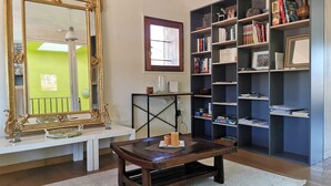 A room with a large ornate mirror, a wooden coffee table with candles, a bookshelf filled with books, and a window with a view of a green building.