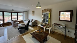 A spacious living room with a sofa, armchairs, a coffee table, and a large mirror. There are multiple windows providing natural light and a view of the outside.