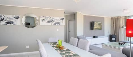 A modern living room with a dining area, grey sofas, a glass coffee table, and artwork on the walls.