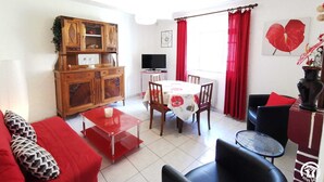 A living room with a red sofa, a wooden cabinet, a dining table with chairs, a TV, and a window with red curtains.
