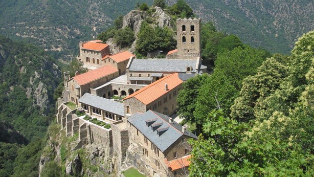 A historic building with multiple structures, including a tower, situated on a hillside with a mountainous backdrop.