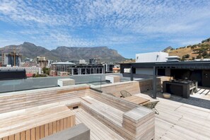 A rooftop terrace with wooden decking, seating areas, and a view of a cityscape and mountains.