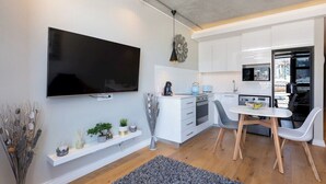 Una cocina moderna con armarios blancos, un televisor de pantalla plana, una zona de comedor con mesa y sillas, y vistas a la ciudad a través de una ventana.