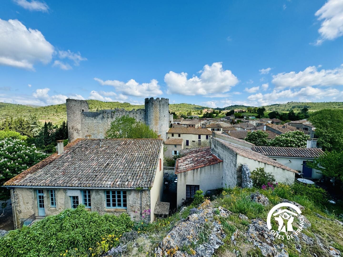 A historic stone building with a tiled roof, surrounded by lush greenery and a clear blue sky.