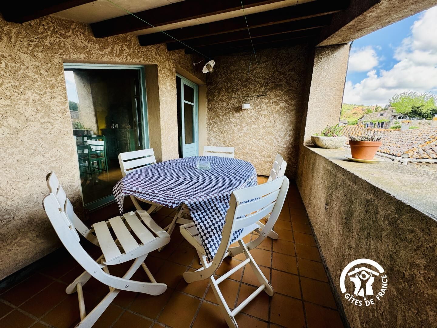 A round table with a checkered tablecloth, surrounded by white chairs, on a balcony with stone walls and a view of the countryside.