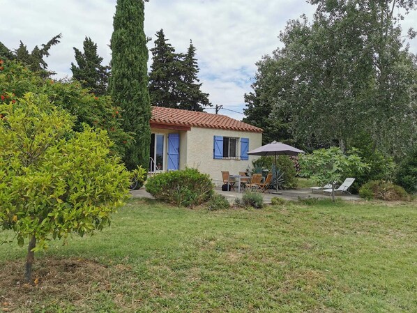 A small house with a tiled roof, a patio with a table and chairs, and a garden with various plants and trees.