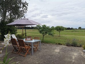 An outdoor patio with a table and chairs, a large umbrella, and a view of a grassy field with trees.