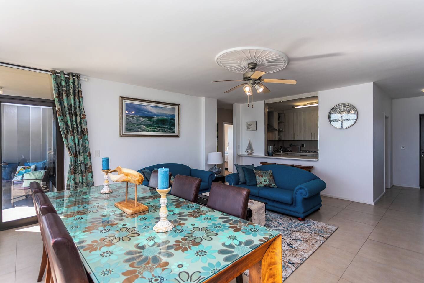 A modern living room with a dining area, a blue sofa, a ceiling fan, and a kitchen in the background.