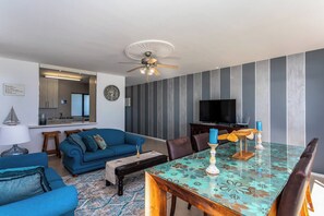 Un salon moderne avec un canapé bleu, une table à manger recouverte d’une nappe à motifs, un ventilateur de plafond et un téléviseur à écran plat placé contre un mur à rayures verticales.