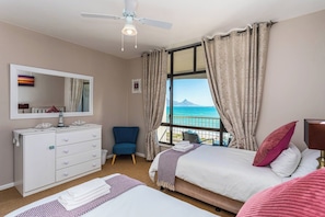 A hotel room with a bed, a chair, a dresser, and a large window with a view of the sea.