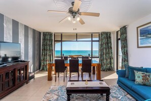 A living room with a television, a dining table with chairs, a sofa, a ceiling fan, and a view of the sea through large windows.