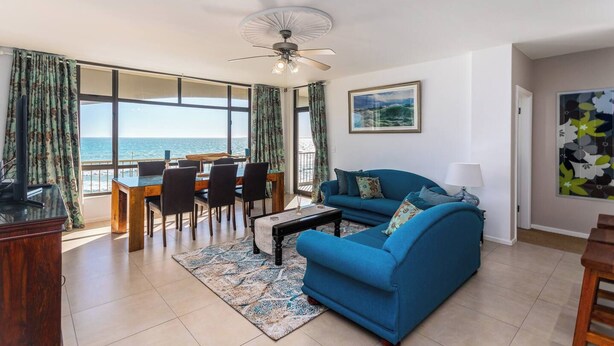 A living room with a blue sofa, a dining table with chairs, a ceiling fan, and a view of the ocean through large windows. - Beachfront 3br | Ocean Views & bbq balcony by ctha (Cape Town)