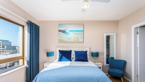 A hotel room with a bed, a chair, a painting, a window, and a ceiling fan. - Beachfront 3br | Ocean Views & bbq balcony by ctha (Cape Town)