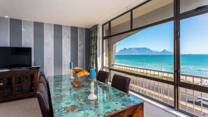 A room with a large window, a television, a table with chairs, and a view of the ocean. - Beachfront 3br | Ocean Views & bbq balcony by ctha (Cape Town)