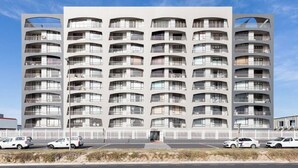 A multi-story hotel with balconies, a white fence, and cars parked in front. - Beachfront 3br | Ocean Views & bbq balcony by ctha (Cape Town)