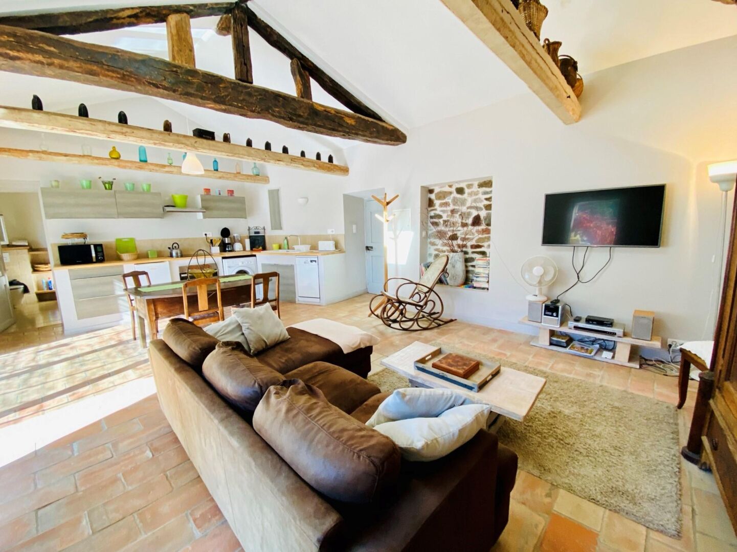A modern living room with a brown leather sofa, a dining area, a kitchen with white appliances, and a stone wall.