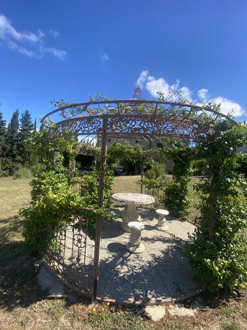Un jardín con una pérgola, bancos de piedra y un cielo azul despejado.