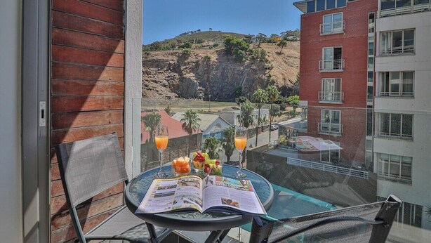 Un balcón con una mesa lista para dos personas, que incluye un menú, fruta y dos vasos de zumo de naranja. Se observa una piscina y un edificio con fachada de ladrillo rojo.