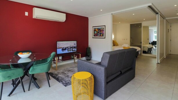 Un salon moderne avec un mur d’accent rouge, un téléviseur à écran plat, un coin repas avec une table en verre et des chaises vertes, et un canapé gris.